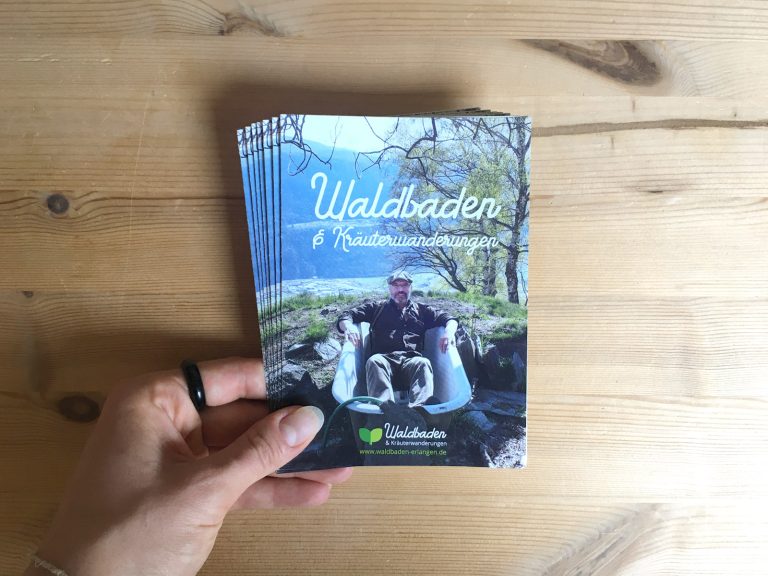 Ein Stoß Waldbaden Flyer der in einer weiblichen linken Hand gehalten wird. Der Hintergrund ist aus Holz.